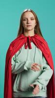 Vertical Smiling teenager portraying superhero wearing red cape, isolated over studio background. Portrait of cheerful heroic person posing as hero in costume with mantle, camera B video