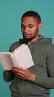 Vertical Man laughing while reading funny book, entertained by engrossing story. Joyful person having fun, being amused by literature novel during leisure time in studio background, camera B video