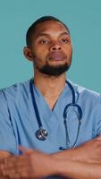 Vertical Portrait of serious professional male nurse standing with arms folded, studio background. Stern healthcare worker wearing protective clinical scrubs to prevent spreading of diseases, camera A video