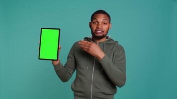 Happy influencer holding isolated screen tablet, doing recommendation, talking with audience. Cheerful person pointing towards chroma key digital device, isolated over studio background, camera B video