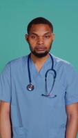Vertical Portrait of serious professional male nurse standing with arms folded, studio background. Stern healthcare worker wearing protective clinical scrubs to prevent spreading of diseases, camera B video