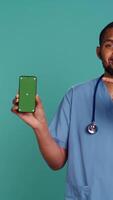 Vertical Portrait of jolly male nurse pointing finger towards green screen smartphone. Upbeat hospital employee holding chroma key phone, isolated over blue studio background, camera B video