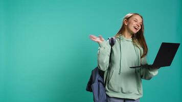 Happy woman saluting friends during videoconference meeting using laptop, studio backdrop. Girl waving hand, greeting mates during internet videocall on notebook, camera B video