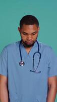Vertical Portrait of joyous african american caretaker standing with arms folded, having positive emotion, studio backdrop. Merry healthcare professional grinning, wearing clinical workwear, camera B video