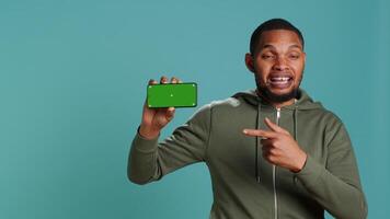 Portrait of upbeat content creator doing influencer marketing using green screen mobile phone. Cheerful person holding chroma key cellphone device, isolated over studio backdrop, camera A video