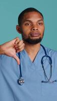 Vertical Sad african american male nurse showing thumbs down signs while at work, isolated over studio backdrop. Unhappy healthcare employee wearing scrubs doing disapproval gestures, camera A video