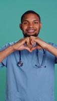 Vertical Portrait of cheerful healthcare professional doing heart symbol shape with hands. Licensed nurse showing love gesturing while at work, isolated over blue studio background, camera B video