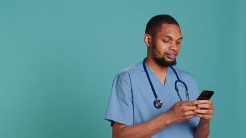 Male nurse using mobile phone to scroll on social media feed during hospital job shift break. Healthcare worker using smartphone to text friends, studio backdrop, camera A video