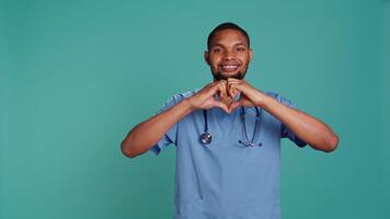 Portrait of cheerful healthcare professional doing heart symbol shape with hands. Licensed nurse showing love gesturing while at work, isolated over blue studio background, camera B video