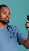 Vertical Male nurse in videocall meeting using mobile phone, greeting people. Healthcare expert in online telemedicine call with patients using smartphone, isolated over studio background, camera A video