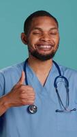 Vertical Cheerful male nurse showing thumbs up signs while at work, isolated over studio background. Friendly healthcare professional smiling and doing approval gestures, camera A video