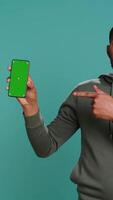 Vertical Portrait of african american man doing influencer marketing using green screen phone, studio background. BIPOC person holding empty copy space mockup smartphone device, camera B video