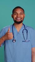 Vertical Cheerful male nurse showing thumbs up signs while at work, isolated over studio background. Friendly healthcare professional smiling and doing approval gestures, camera B video