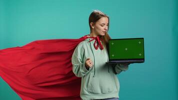 Young girl wearing superhero costume presenting and talking about green screen laptop, studio background. Teenager dressed as comic book hero advertising chroma key notebook, camera B video