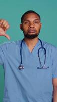 Vertical Sad african american male nurse showing thumbs down signs while at work, isolated over studio backdrop. Unhappy healthcare employee wearing scrubs doing disapproval gestures, camera B video