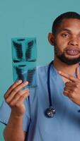 Vertical Healthcare professional informing patient of radiology x ray scan results, studio backdrop. African american doctor presenting radiography and providing injury treatment, camera A video