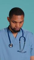 Vertical Portrait of joyous african american caretaker standing with arms folded, having positive emotion, studio backdrop. Merry healthcare professional grinning, wearing clinical workwear, camera A video
