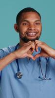 Vertical Portrait of cheerful healthcare professional doing heart symbol shape with hands. Licensed nurse showing love gesturing while at work, isolated over blue studio background, camera A video