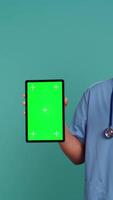 Vertical Portrait of male nurse showing medical instructions on green screen tablet. Hospital employee holding chroma key device, isolated over blue studio background, camera B video