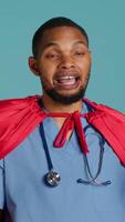 Vertical Male nurse portraying superhero showing courage and strength fighting diseases, studio backdrop. Portrait of african american doctor posing as hero in costume, holding empowering speech, camera A video