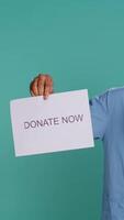 Vertical Portrait of smiling medic holding donate now sign, inviting people to provide aid for those in need, studio background. Healthcare employee requesting philanthropic donations for patients, camera B video