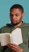 Vertical Man laughing while reading funny book, entertained by engrossing story. Joyful person having fun, being amused by literature novel during leisure time in studio background, camera A video