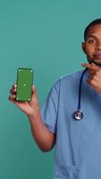 Vertical Portrait of joyful african american male nurse pointing finger towards green screen cellphone. Smiling healthcare professional holding mockup mobile phone, studio background, camera B video