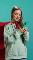 Vertical Young girl wearing superhero costume texting friends using smartphone, studio backdrop. Woman dressed as comic book hero entertained by chatting with mates using mobile phone, camera B video