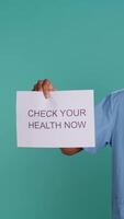 Vertical Portrait of upbeat nurse smiling, holding sign urging people to do doctor appointments, studio background. African american healthcare worker talks about importance of preventive healthcare, camera B video