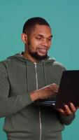 Vertical Smiling man scrolling on laptop, using internet and credit card to pay for products. African american person happily browsing online shopping websites, isolated over studio background, camera B video