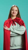 Vertical Radiant woman grinning while portraying confident superhero wearing red cape, isolated over studio backdrop. Portrait of jolly heroic person posing as powerful hero character in costume, camera B video