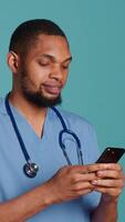 Vertical Male nurse using mobile phone to scroll on social media feed during hospital job shift break. Healthcare worker using smartphone to text friends, studio backdrop, camera A video