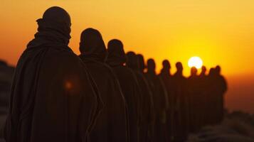 As the sun sets on the horizon a line of figures in dark robes stands in a row faces concealed. They are participating in a . photo