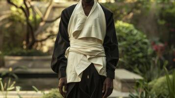 In a tranquil garden setting a regal black man stands tall clad in a flowing ensemble that evokes the flowing forms of organic structures. The way the fabric embraces his curves and photo