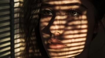 The sunlight filters in through the slatted blinds of a dimly lit room casting shadows across the face of a woman with strikingly bold features. Her expression is one of quiet photo