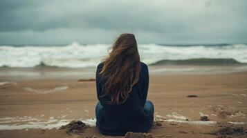 A woman rests head on knees as sits on a secluded beach back facing the camera. The crashing waves provide a calming . photo