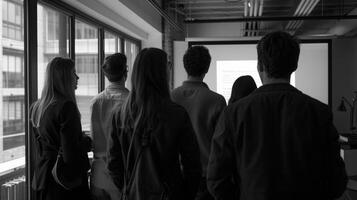 The group of professionals stand around a large projector backs turned to the camera as they review the slides for client . photo