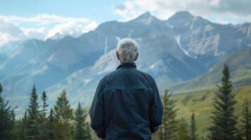 Against a backdrop of mountains and trees an elderly man stands facing away from the camera gazing out at the vast landscape before . photo