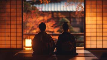 The backs of two individuals dressed in traditional Japanese garb are silhouetted against the warm inviting glow of a traditional . photo