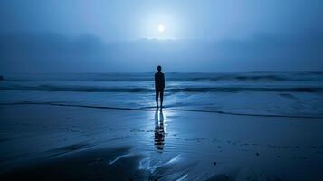 A figure stands alone on a deserted beach back to the camera facing the open sea. body is outlined by the soft glow of . photo