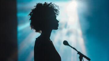 A silhouette of an interpreter on the stage standing beside a speaker and relaying their words to a diverse audience. The lights are dim but their presence is prominent representing photo