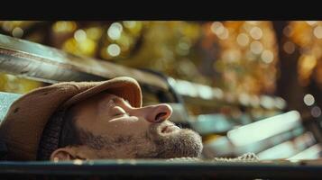 A man sleeps on a bench in a park his hat pulled down over his face and his body resting against the backrest. Despite his unconventional sleeping spot his face is peaceful and his photo