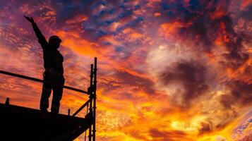 The silhouette of a construction worker appears to be reaching out to the sky as he stands on a scaffold against a vibrant sunset photo