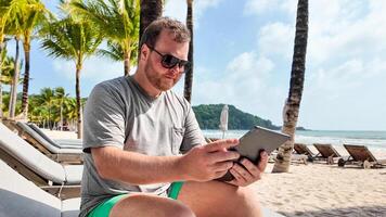 caucásico hombre utilizando un tableta en un tropical playa con palma árboles, encarnando ocio tecnología utilizar y verano vacaciones foto