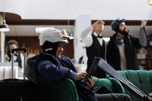 Female tourist seated in cozy hotel lobby surfing the net on smart tablet. Asian lady with device dressed in ski gear and ski goggles resting on forehead hints at upcoming adventure on winter slopes. photo
