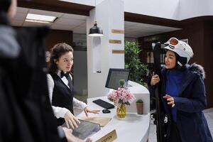 Side-view shot of concierge providing excellent service to asian woman grasping her ski equipment. Staff catering to every need of winter resort guest in snow clothing creating a joyful atmosphere. photo