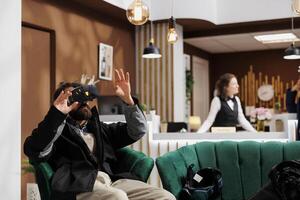 Male tourist wearing winter jacket is using a vr headset in lounge area of ski mountain resort. While waiting in hotel lobby man with beard utilizes vr goggles for research on wintersports activity. photo