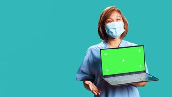 Close up handheld camera shot of happy clinic worker showing instructions on laptop chroma key green screen. Nurse isolated over studio background presenting healthcare tape on gadget photo