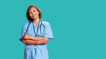 Joyful experienced asian nurse folding arms, smiling while looking at camera. Portrait of happy healthcare expert wearing protective scrubs, isolated over blue studio background photo
