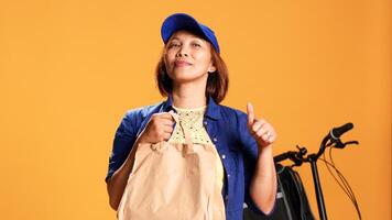 Slow motion close up portrait shot of courier waiting for customer to pick up food order. Bicycle rider delivering meal bag to client, showing approval gesturing, isolated over studio background photo